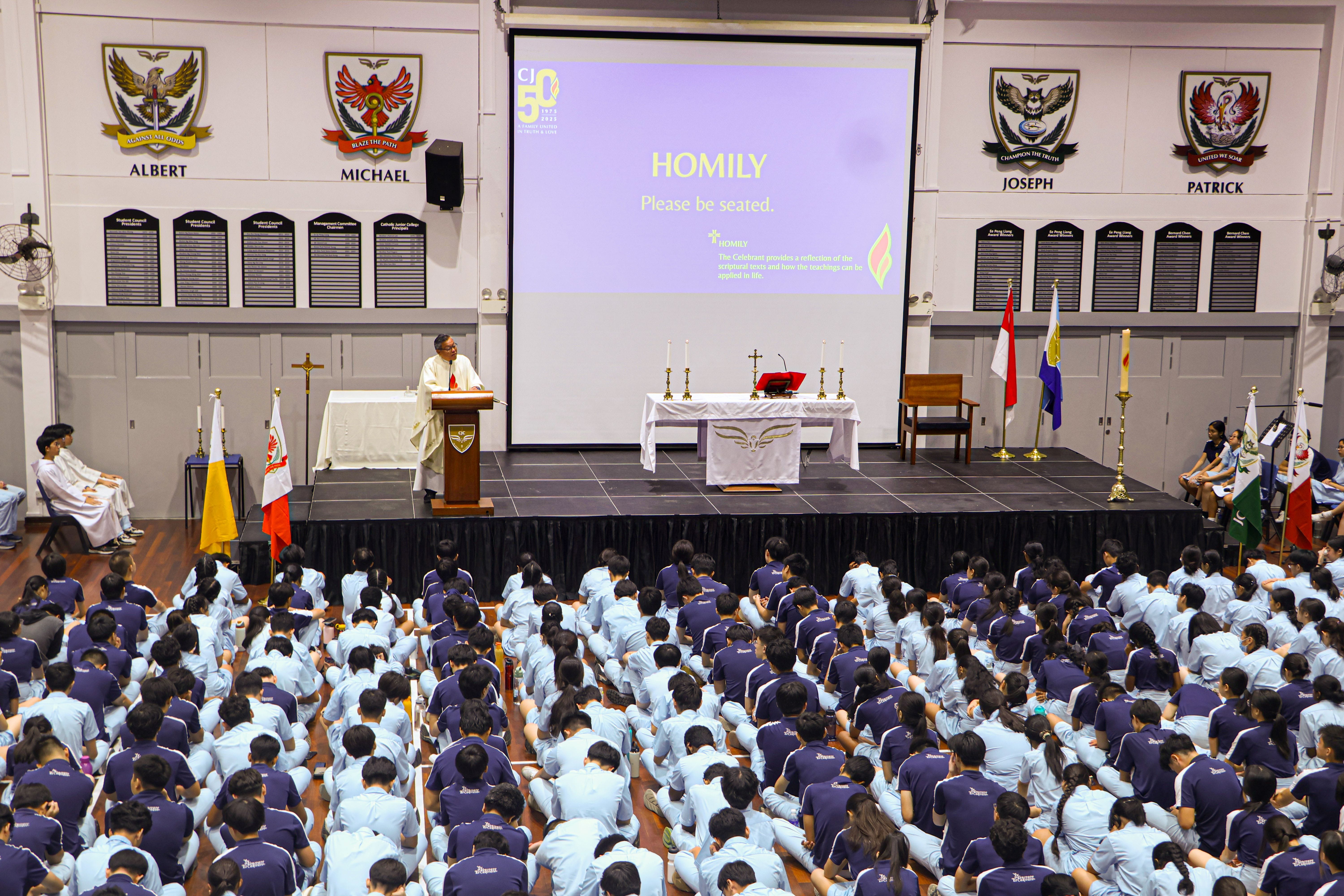 A priest delivers a homily at a podium to uniformed students seated on a hall floor, with a projected slide reading "HOMILY."